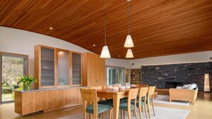 A modern kitchen with wooden cabinets, a dining area with a table and chairs, and a stone fireplace. - Contemporary Berkshire Home with Views of Taconic Golf Course (Williamstown)