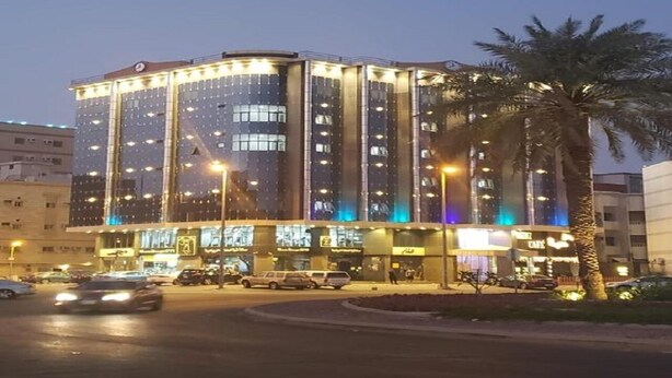 Un hotel a più piani dal design moderno, con un ingresso ben visibile dotato di insegne illuminate, un'area di parcheggio antistante e una palma nelle vicinanze.