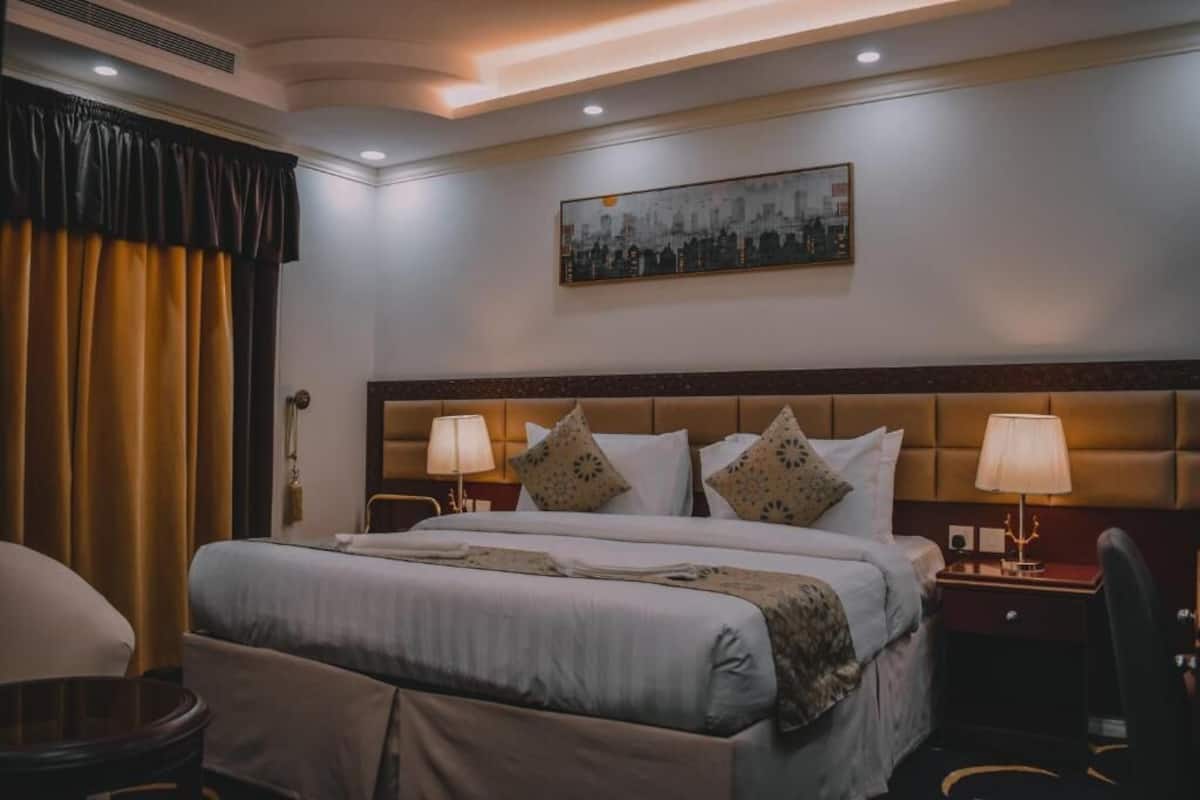 A hotel room with a large bed, two bedside tables with lamps, a headboard, a painting on the wall, and a window with curtains.