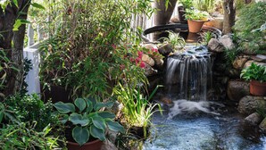 Ein Garten mit einem kleinen Wasserfall, Blumentöpfen und einem weißen Zaun.
