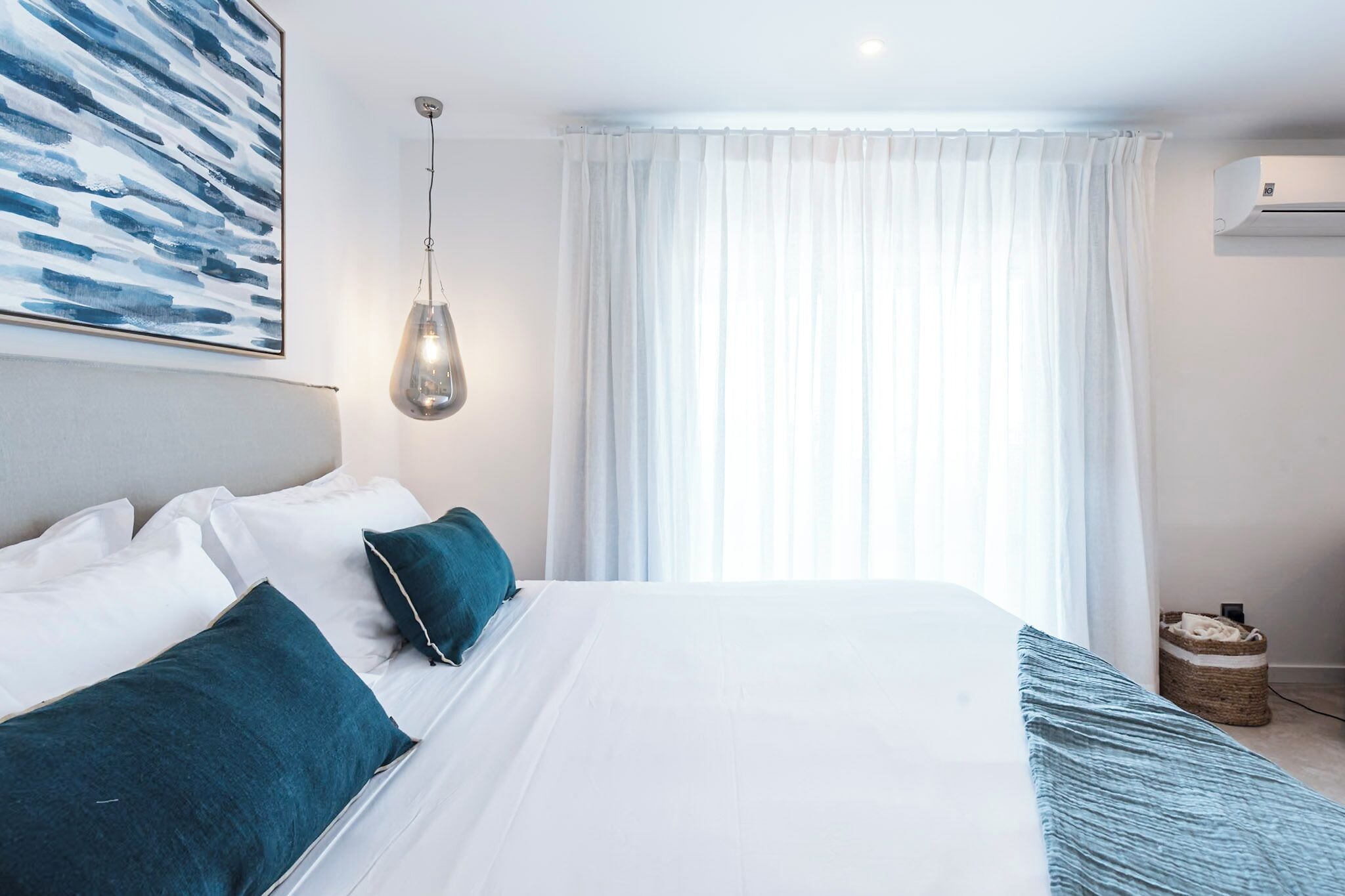 A neatly made bed with white and teal bedding, a hanging light fixture, and a large window with sheer curtains.