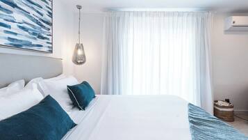A neatly made bed with white and teal bedding, a hanging light fixture, and a large window with sheer curtains.