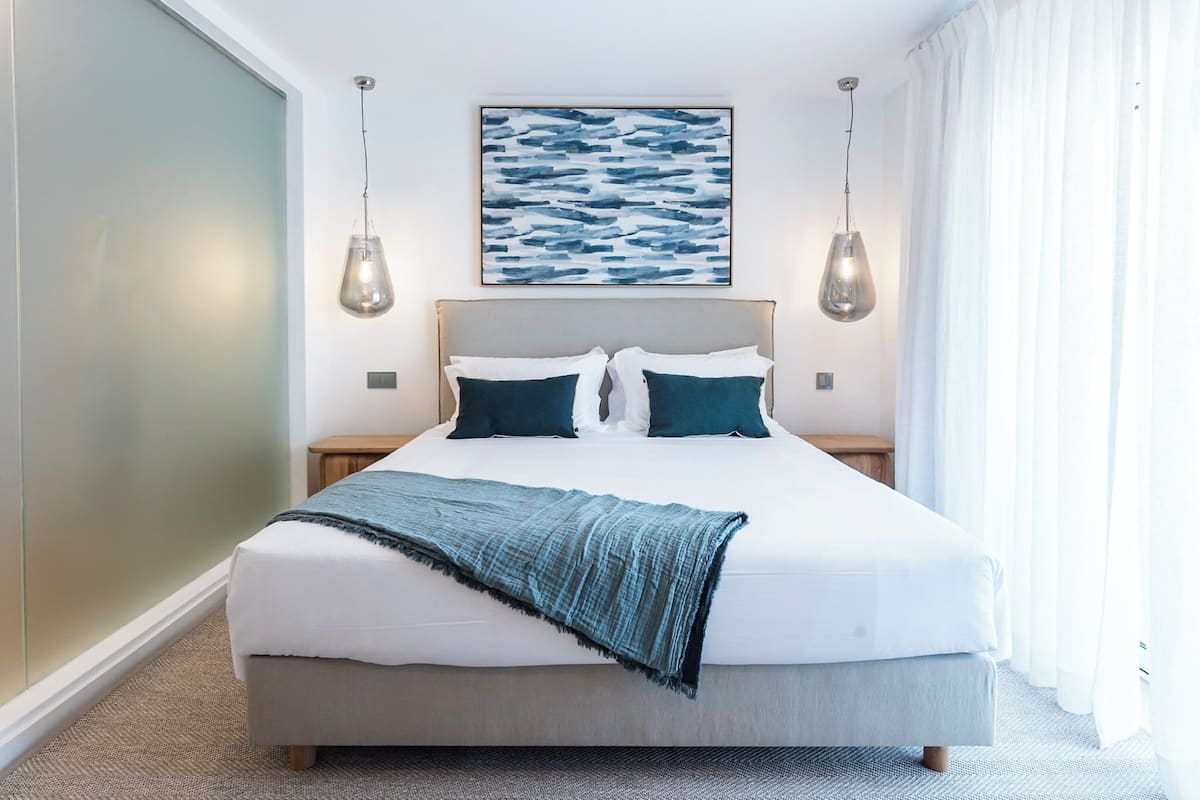 Une chambre d’hôtel moderne dotée d’un grand lit, de deux tables de chevet, d’une œuvre d’art accrochée au mur et d’une grande fenêtre avec des rideaux.