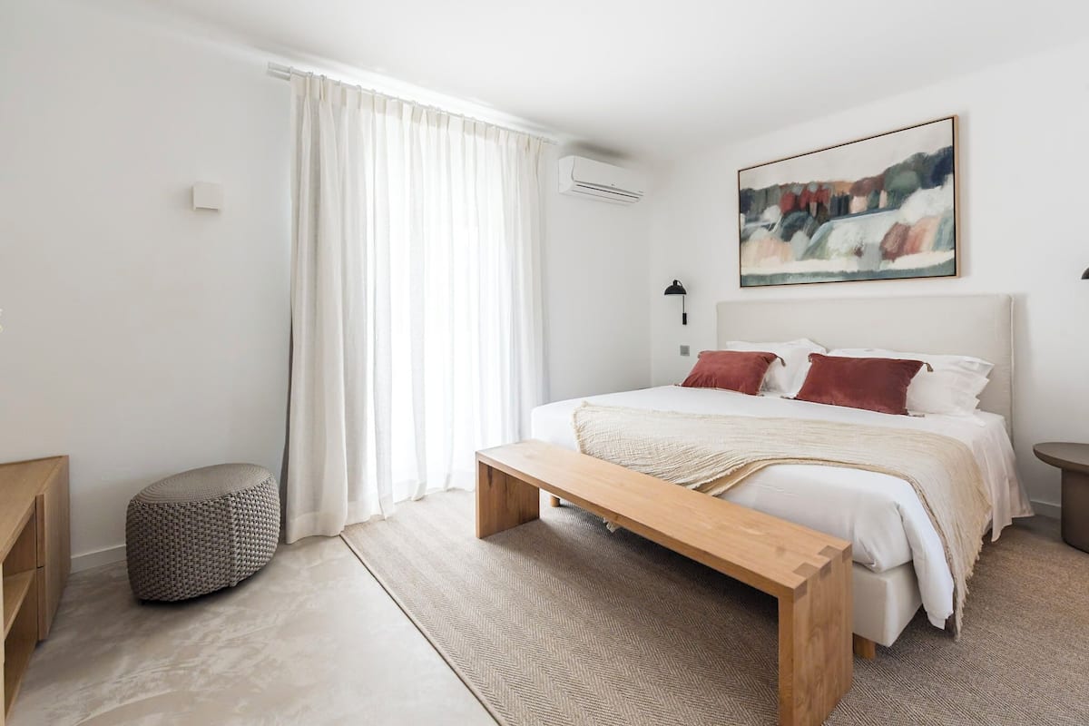 Une chambre moderne avec un grand lit, un banc, une table de chevet et une œuvre d’art suspendue au mur.