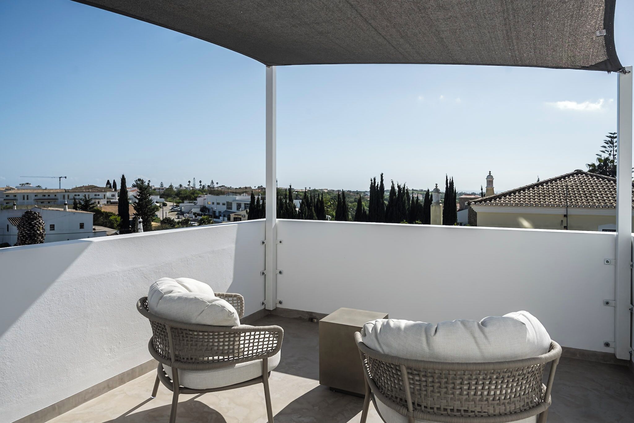 Un balcon avec deux fauteuils en osier et une petite table, d’où l’on peut admirer une vue sur la ville, avec ses bâtiments et ses arbres.