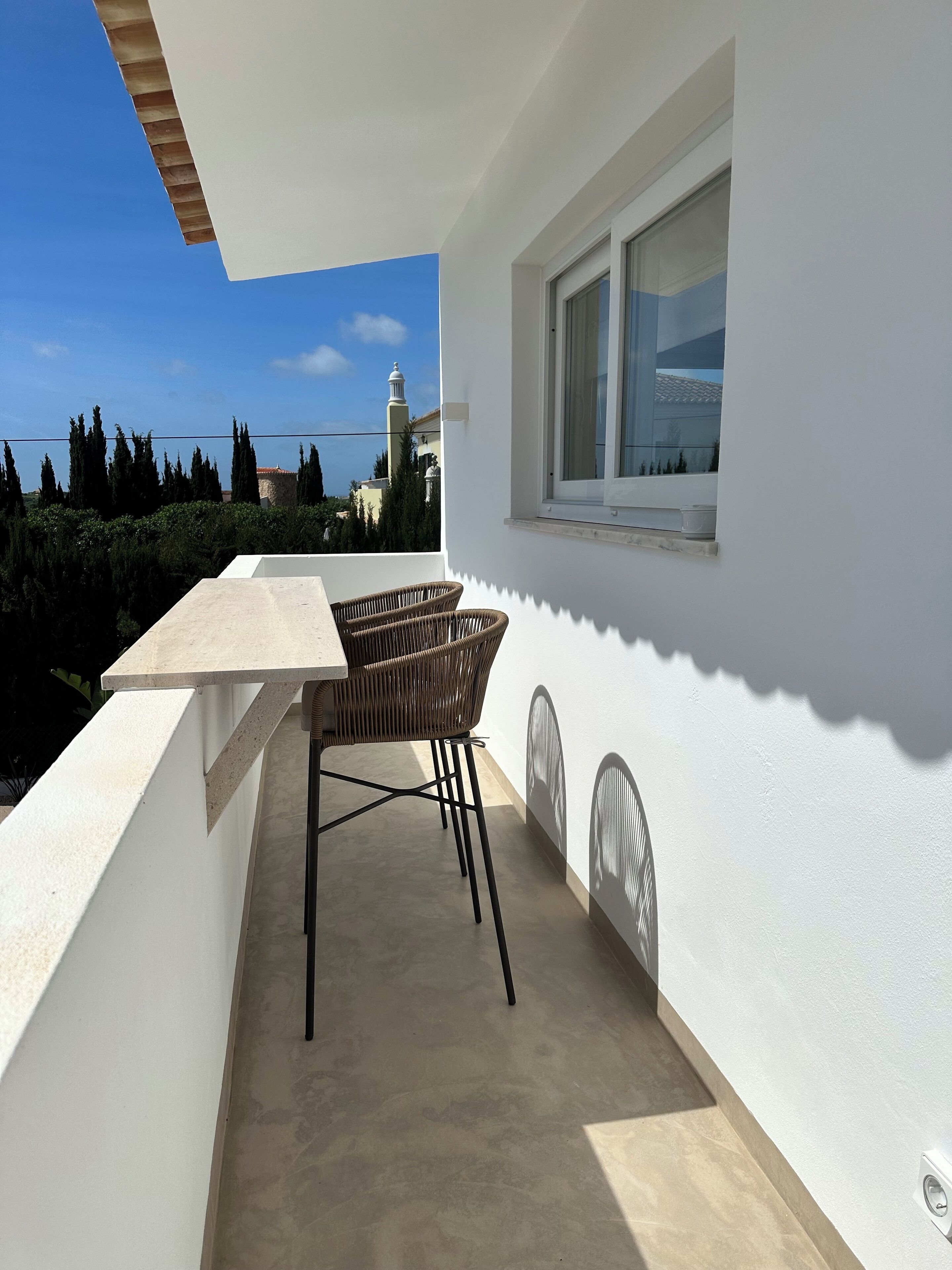 Ein Balkon mit Tisch und Stühlen, ein Fenster und Blick auf ein weit entferntes Gebäude und Bäume.