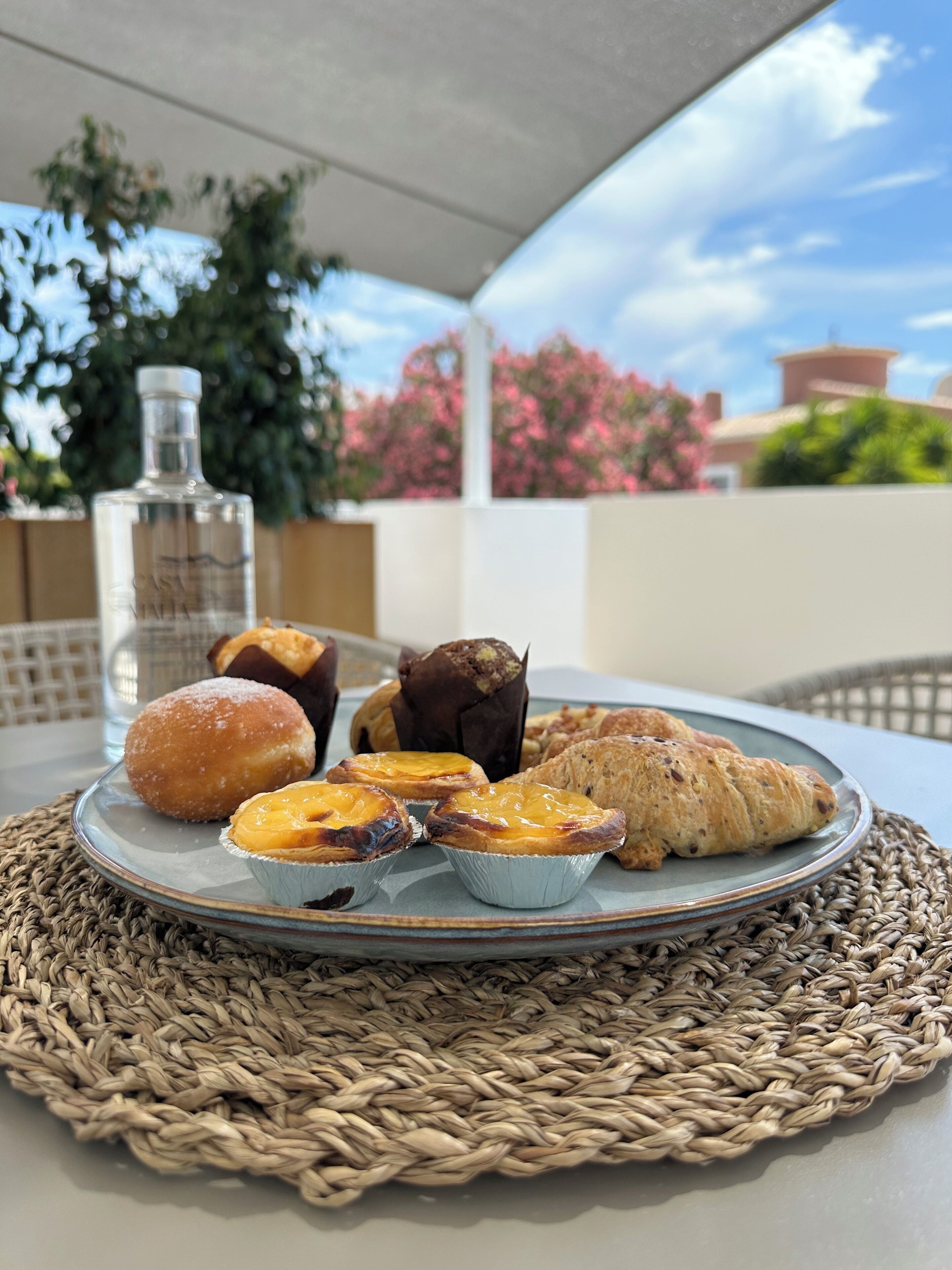 A table with a woven placemat, a plate of pastries including a croissant, a muffin, and a tart, and a bottle of water.