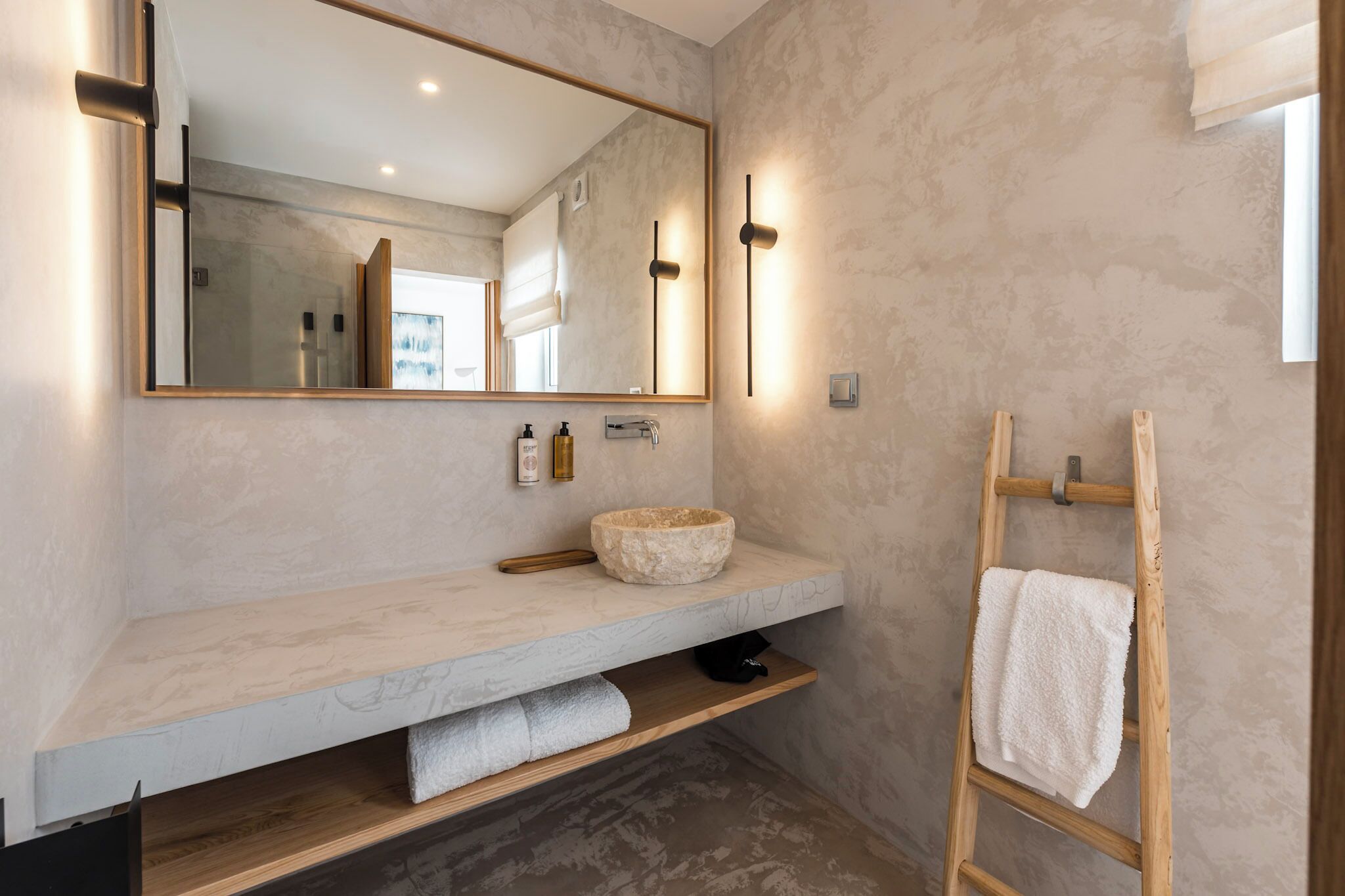 Un baño moderno con un espejo grande, un lavabo de piedra, un estante de madera y un perchero para toallas.