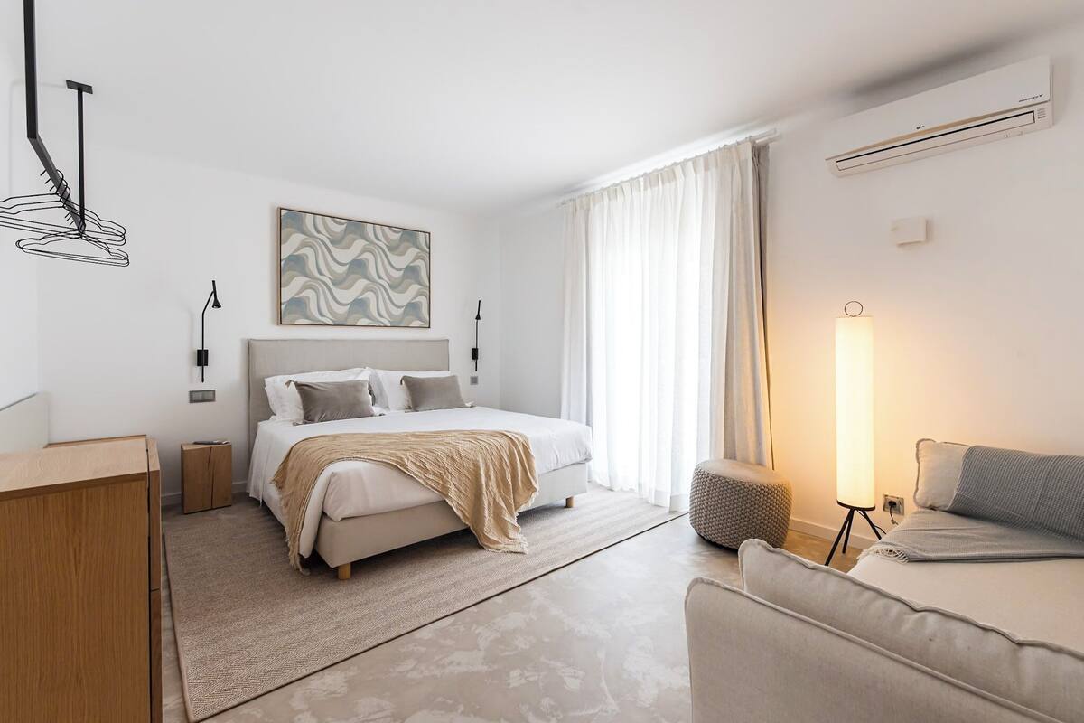 Une chambre moderne avec un lit, des tables de chevet, une lampe de chevet, un climatiseur mural et une grande fenêtre avec des rideaux.