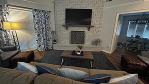 Un salon doté d’une cheminée en briques, d’un téléviseur à écran plat fixé au-dessus, d’un canapé avec des coussins, d’une table basse et d’un rideau à motifs.
