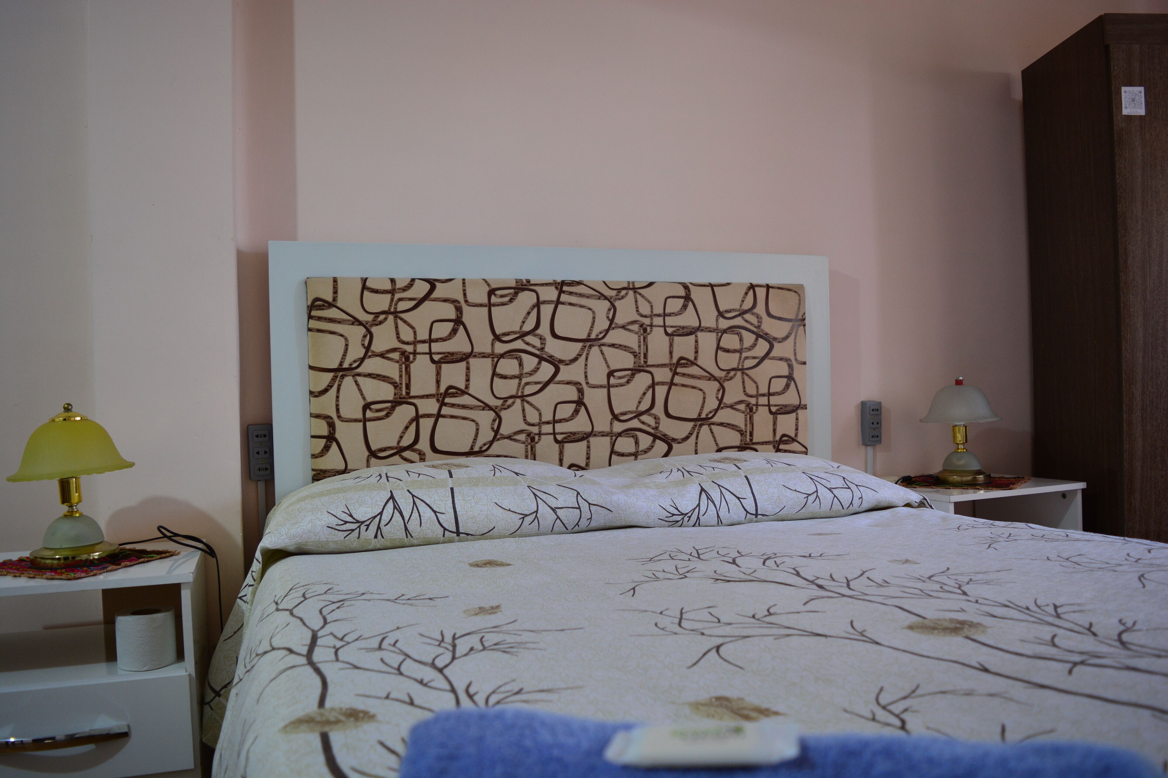 Una cama bien hecha con un cabecero estampado, dos lámparas de noche y una mesita de noche pequeña y blanca con una lámpara.