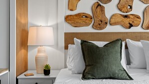 Una cama bien hecha con ropa blanca y un almohadón verde, una lámpara de noche, una pequeña planta en maceta y un cabecero de madera.