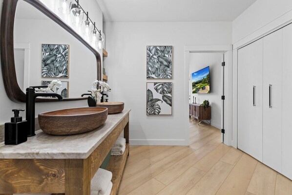 Un baño moderno con un espejo grande, tocador de madera y dos impresiones enmarcadas de hojas en la pared.