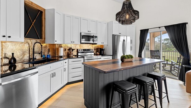 Una cocina moderna con armarios blancos, una isla central con taburetes, electrodomésticos de acero inoxidable y un amplio ventanal con vistas al exterior.