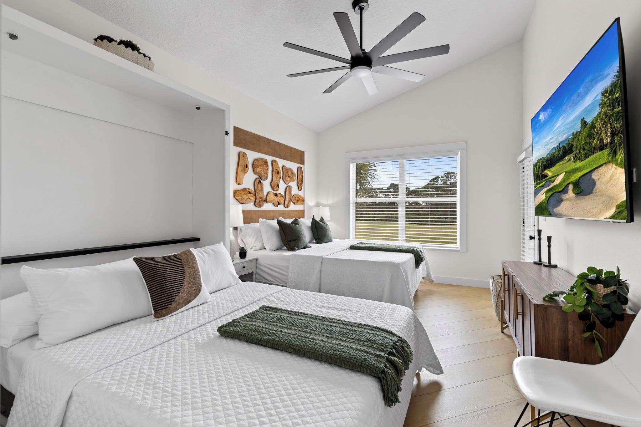A modern bedroom with two beds, a ceiling fan, a large TV, and a window with blinds.