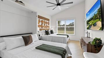 A modern bedroom with two beds, a ceiling fan, a large TV, and a window with blinds.