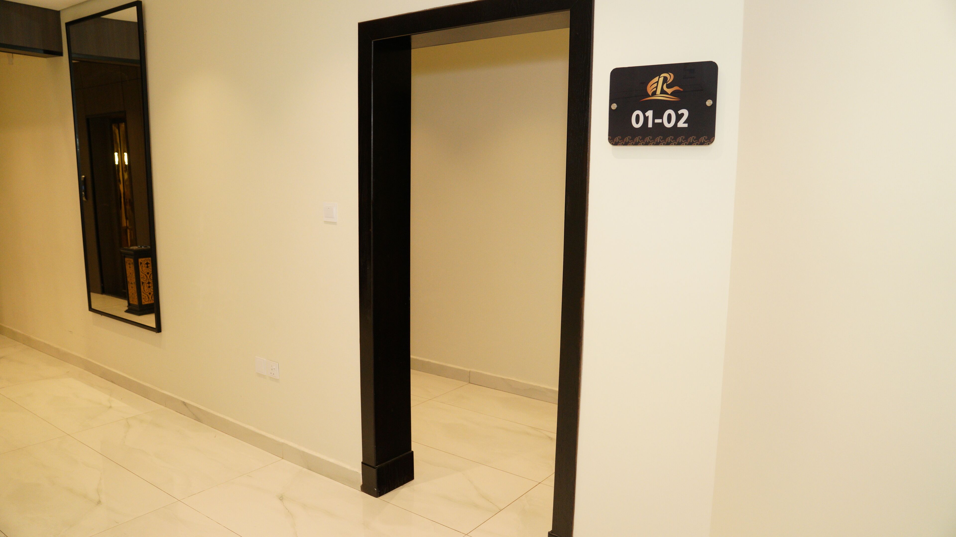 Um corredor com uma porta emoldurada em preto, um espelho e uma placa com '01-02' em uma parede branca.