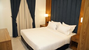 Una cama bien hecha con sábanas blancas, una mesita de noche de madera con una lámpara y un telón oscuro de fondo.