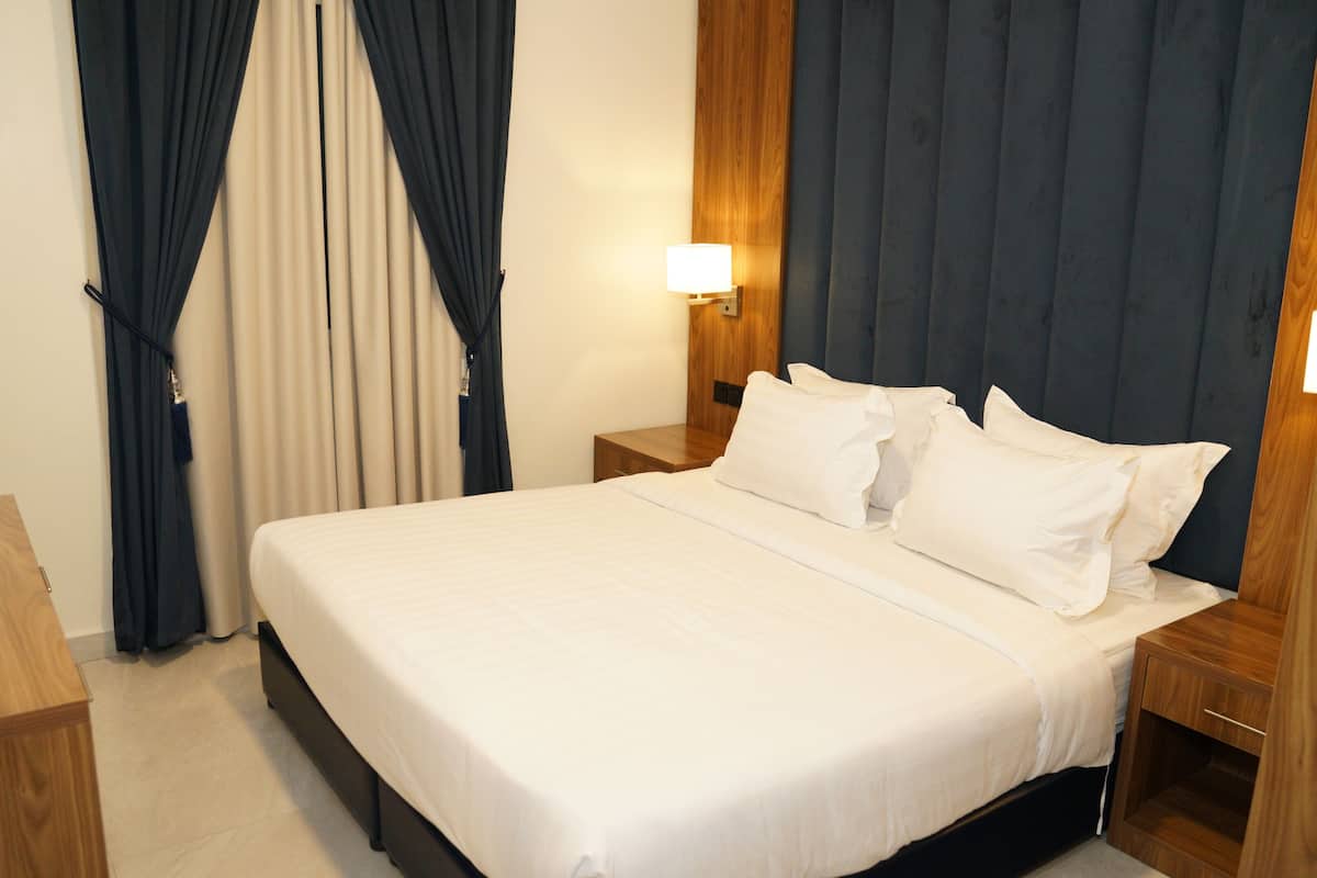Una cama bien hecha con sábanas blancas, una mesita de noche de madera con una lámpara y un telón oscuro de fondo.