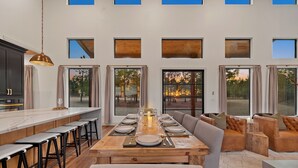 A modern dining area with a long wooden table set for six, surrounded by bar stools and cushioned seating. - Luxury Hilltop, Surreal Views, Pool, Hot Tub, Firepit & 4Kings Bed (Broken Bow)