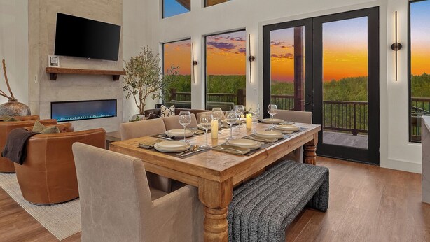 A dining area with a wooden table set for four, a television mounted on the wall, and large windows with a sunset view. - Luxury Hilltop, Surreal Views, Pool, Hot Tub, Firepit & 4Kings Bed (Broken Bow)