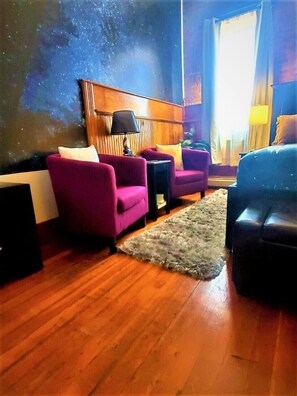 Una habitación con suelo de madera, dos sillones morados, un sofá negro, una alfombra pequeña y una pared con diseño de estrellas.