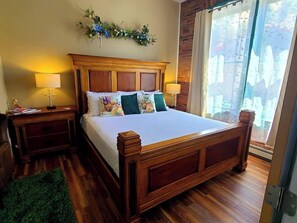 A hotel room with a wooden bed, two bedside tables with lamps, a green rug, and a view of the outdoors through a large window. - Aspen Inn - Spring Suite (Euro King) (Creede)