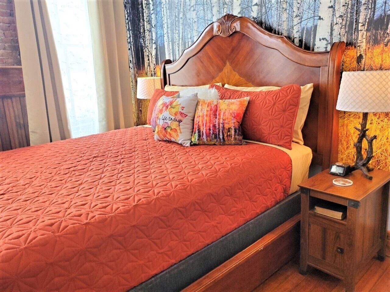 A neatly made bed with a red quilt, multiple pillows, and a wooden headboard. There is a nightstand with a lamp and a small table beside the bed.