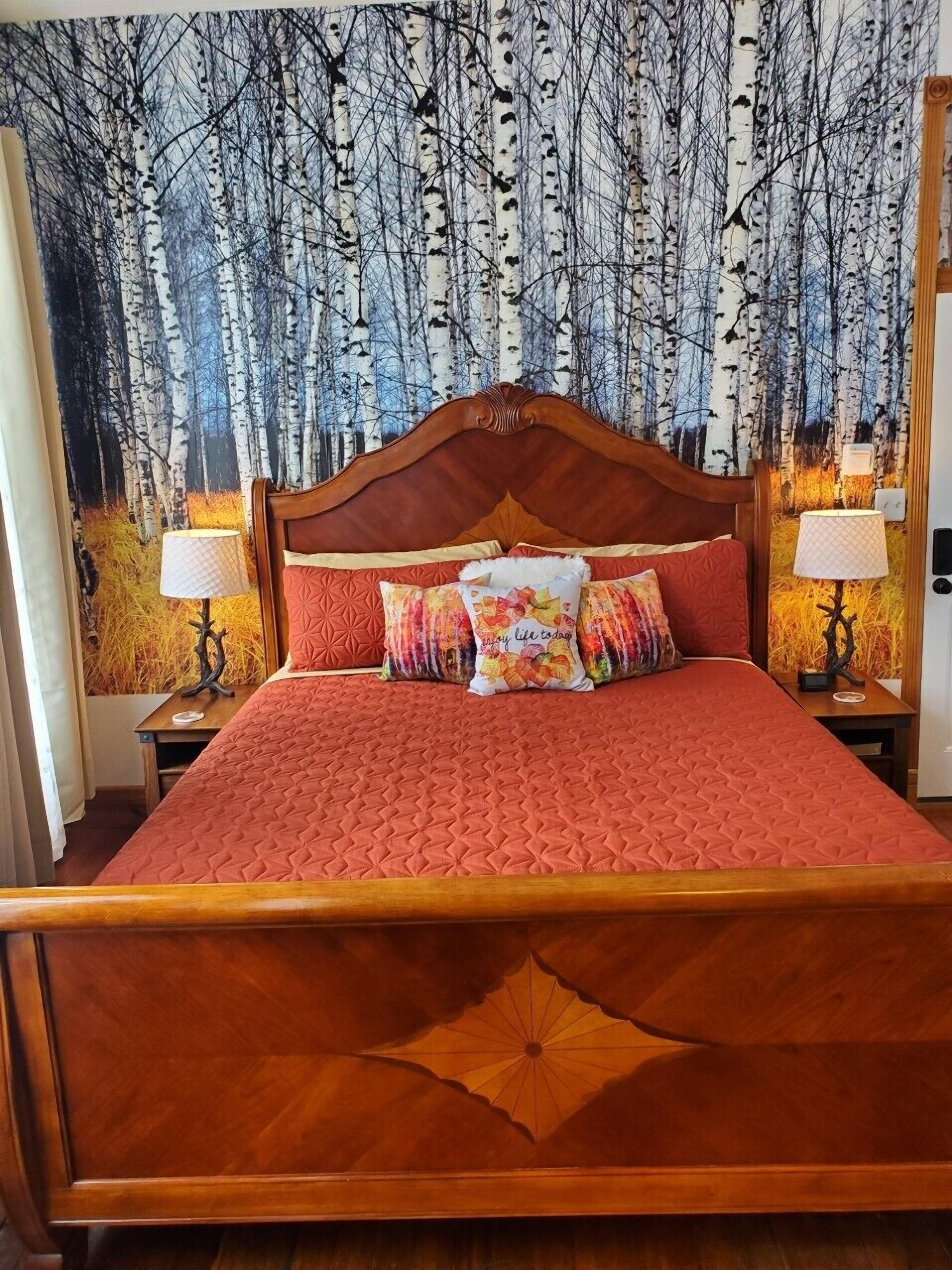 A bed with a wooden headboard, red bedding, and decorative pillows. There are two bedside tables with lamps. The wall behind the bed features a large nature-themed tapestry.
