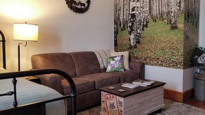 A living room with a brown sofa, a coffee table, a lamp, and a large forest-themed painting on the wall. - Aspen Inn - Summer Suite (Creede)