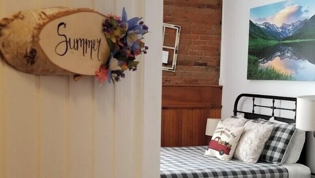 A bedroom with a bed, a dresser, a nightstand, a chair, a painting, and a door with a 'Summer' sign. - Aspen Inn - Summer Suite (Creede)