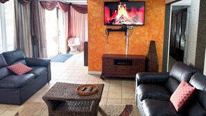 Un salon avec deux canapés en cuir noir, un téléviseur fixé sur un mur orange à texture, et un ventilateur de plafond.