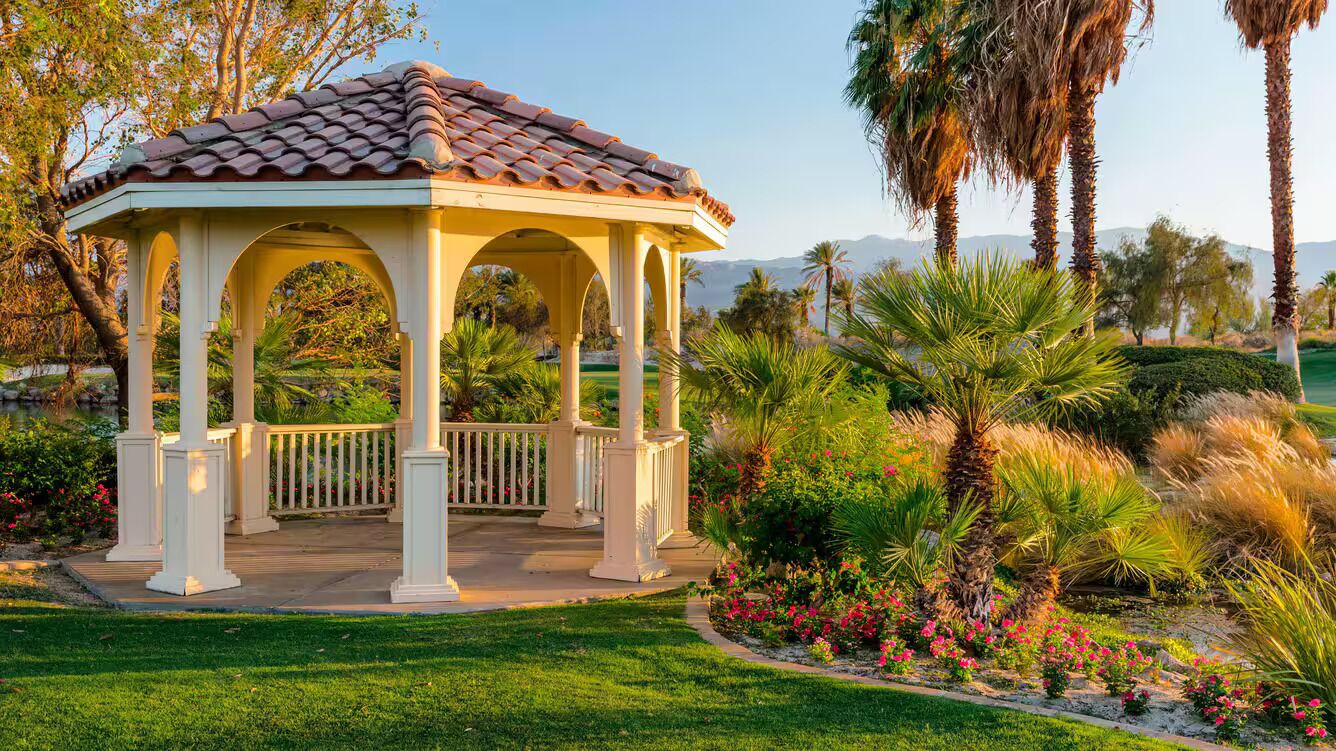 A gazebo with a tiled roof and white columns, surrounded by lush greenery and flowering plants.