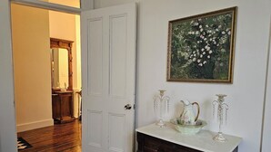 A hallway with a white door, a wooden floor, a mirror, a dresser with drawers, a vase, and a framed painting on the wall.