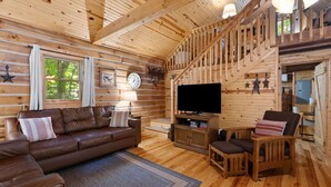 Interior de una cabaña de madera con un sofá de cuero marrón, un televisor y una escalera de madera.