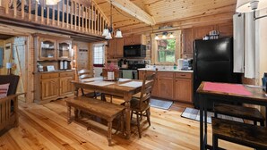 Interior de una cabaña de madera con comedor, cocina y un dormitorio en entrepiso con balcón.