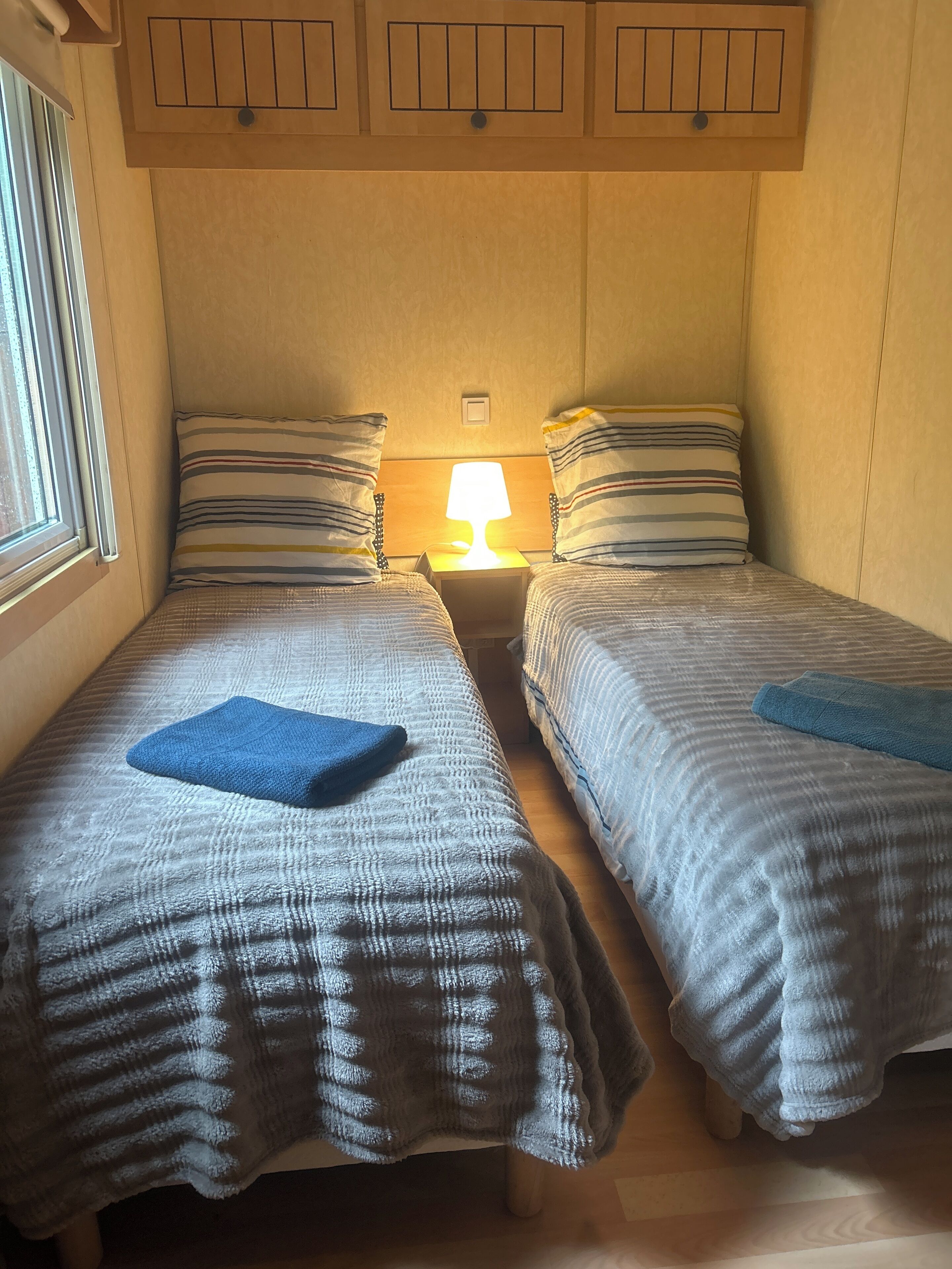 Una habitación pequeña y acogedora con dos camas, cada una con ropa de cama a rayas y una toalla azul. Una lámpara de noche ilumina el espacio, y el diseño es sencillo y funcional.