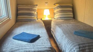 Una habitación pequeña y acogedora con dos camas, cada una con ropa de cama a rayas y una toalla azul. Una lámpara de noche ilumina el espacio, y el diseño es sencillo y funcional.
