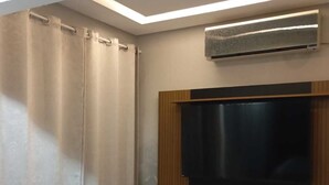 Una habitación con un televisor de pantalla plana grande montado en un panel de madera, un aire acondicionado instalado en el techo y un par de cortinas.