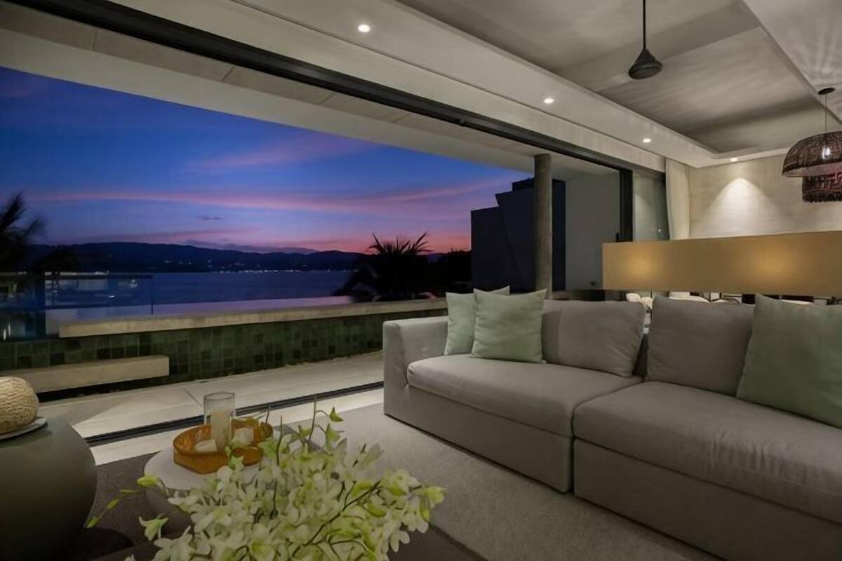 A modern living room with a sofa, a coffee table with a vase of flowers, and a view of a body of water at dusk.
