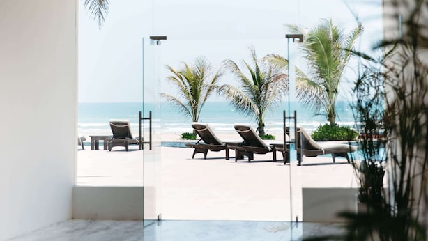 A modern indoor space with a glass wall leading to a beach view, featuring lounge chairs and palm trees.