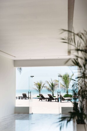 Un espacio interior moderno con una pared de vidrio que da a vista de playa, con sillas de descanso y palmeras.