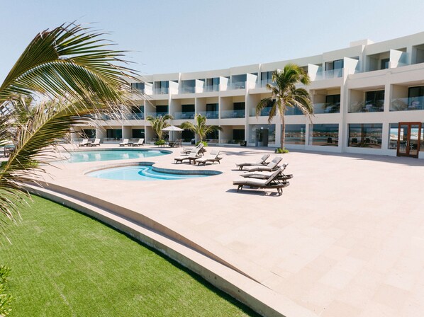 Zona de piscina en un resort con numerosas tumbonas, palmeras y un edificio grande con balcones.
