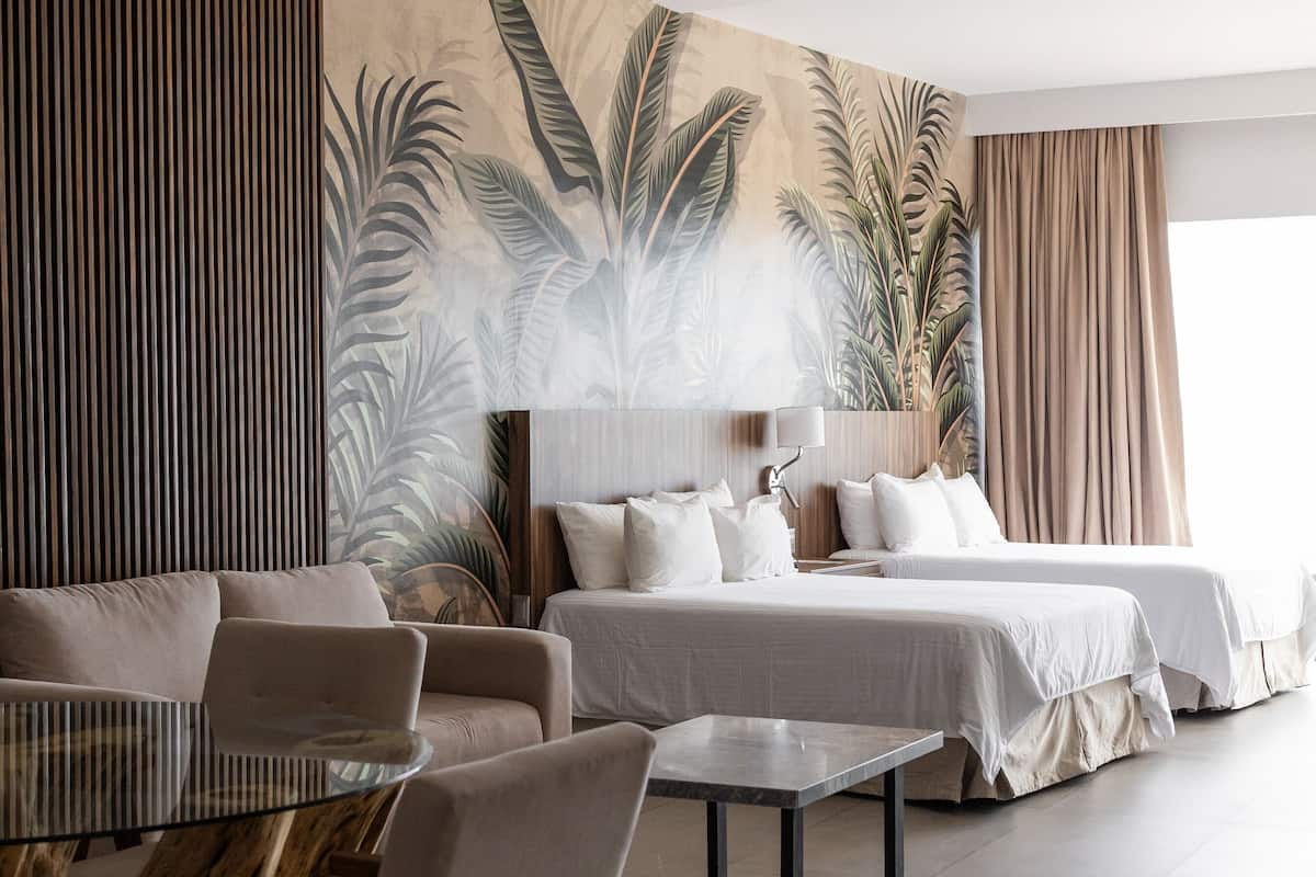 Una habitación de hotel moderna con una cama grande, un sofá, una mesita de vidrio y un mural de hojas tropicales en la pared.