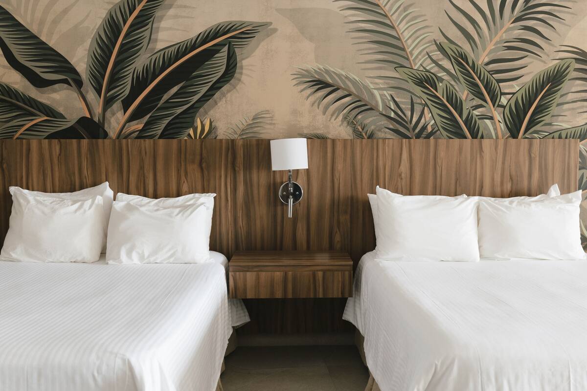 Habitación de hotel con dos camas, cabecera de madera, mesita de noche y pared con estampado de hojas tropicales.