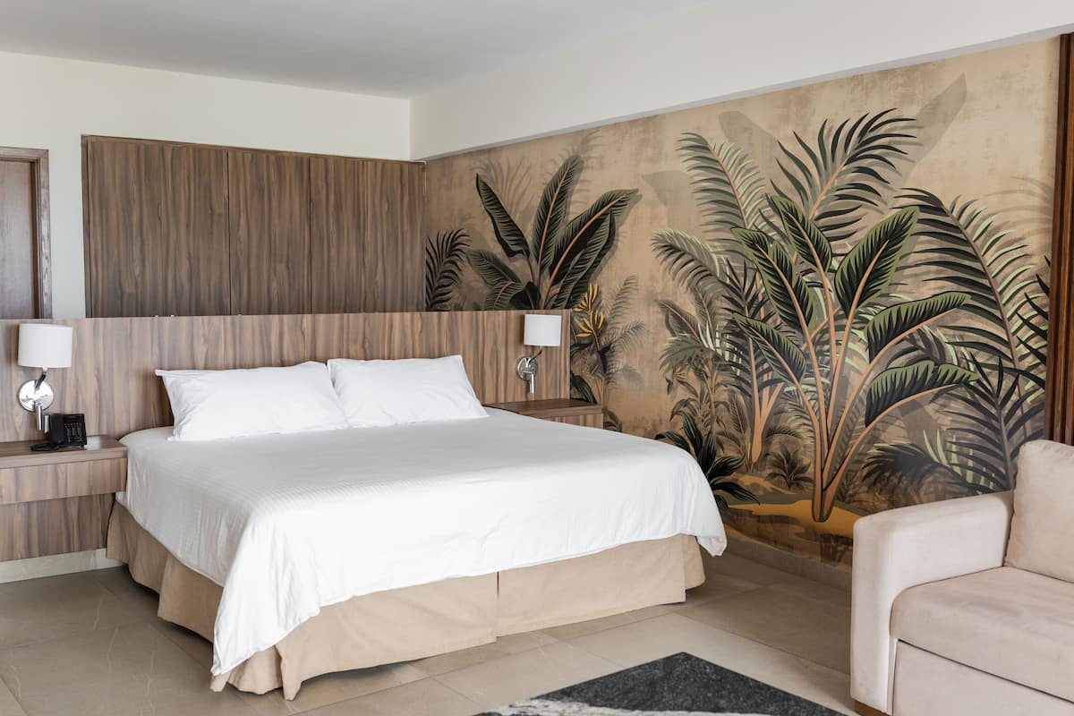 Un dormitorio con una cama grande, una mesita de noche con lámpara, cabecero de madera y un mural de plantas tropicales en la pared.