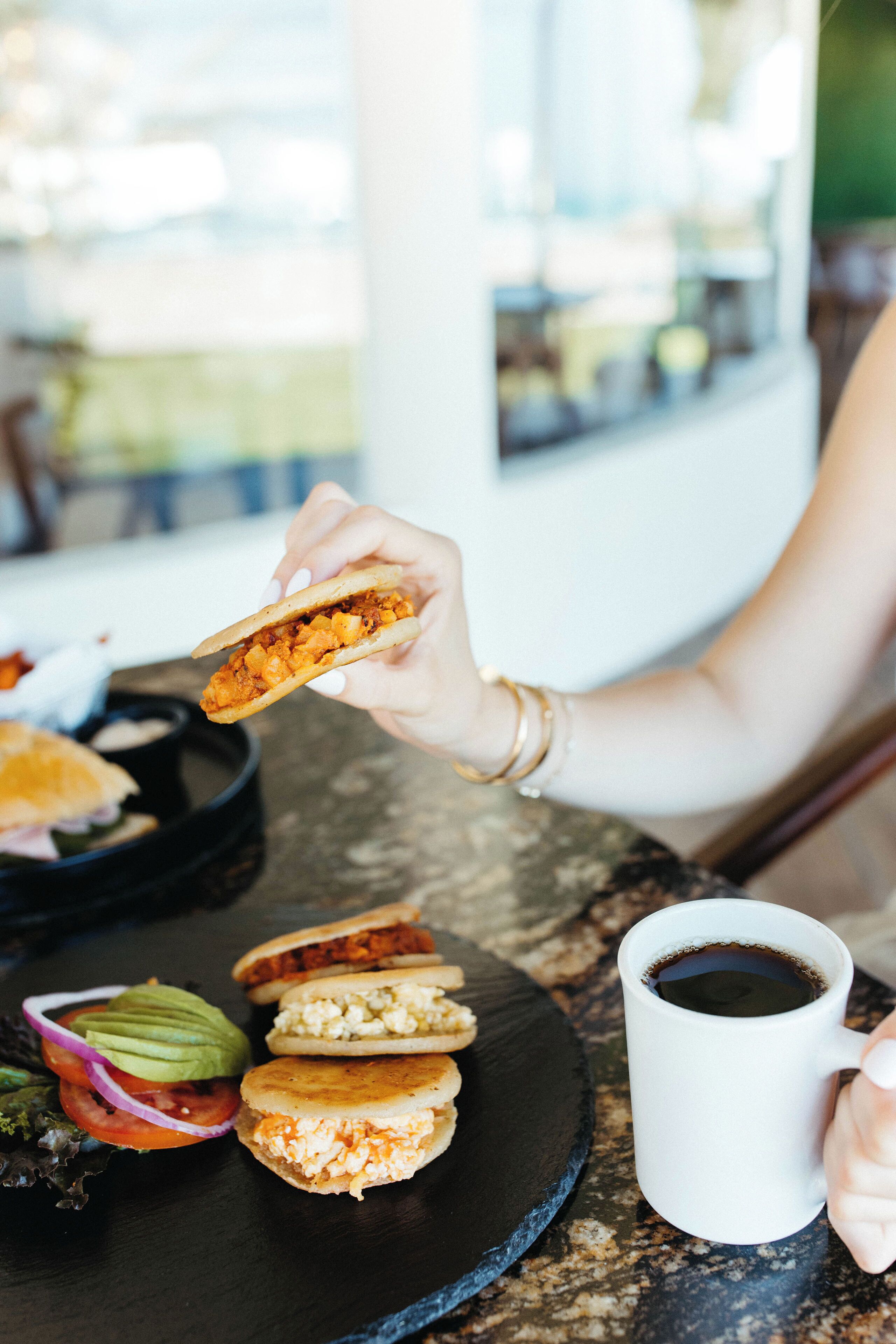 Uma pessoa segurando um sanduíche, com um prato de comida e uma xícara de café sobre a mesa.