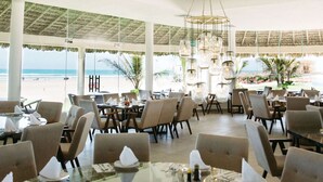 A spacious outdoor dining area with tables and chairs, large windows overlooking the beach, and a thatched roof structure.