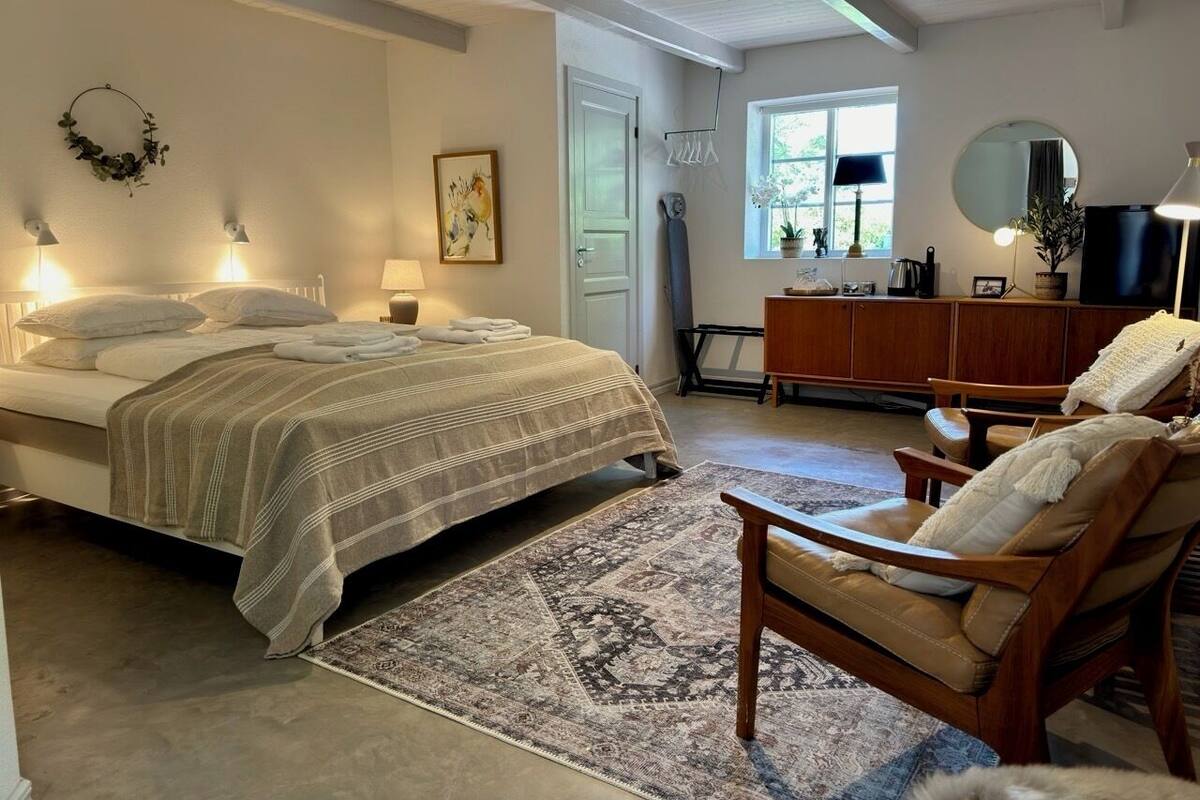 Ein Schlafzimmer mit einem Bett, Nachttischlampen, einer Holzkommode, einem Sessel und einem Fenster mit Blick auf Grünflächen.
