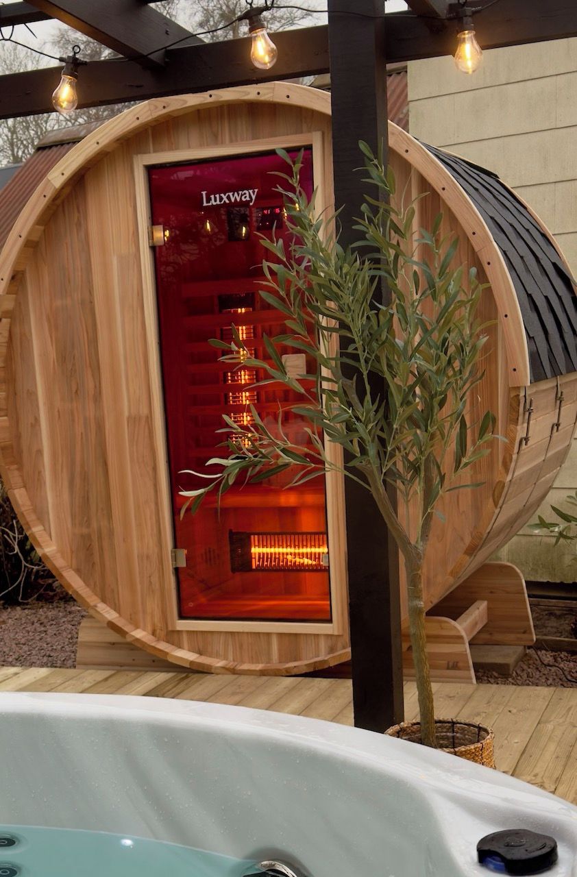 Un sauna de fusta amb una porta vermella que diu "Luxway", un arbre petit i una banyera d'hidromassatge al primer pla.